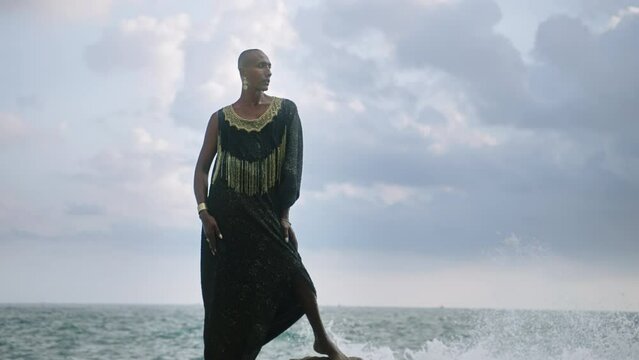 LGBTQIA black person in boutique dress, jewelry stands on rock on stunning ocean coastline skyline. Gender fluid ethnic divine model in luxury outfit fluttering in the wind against dramatic sky.