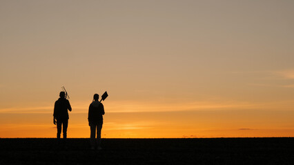 Silhouettes of a couple of farmers with working equipment. In a field at sunset. Back view