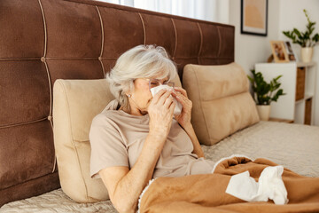 An ill senior woman laying down in bed, sneezing and blowing her nose.