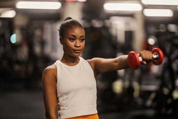 A black sportswoman in shape with dumbbells practicing her shoulders at gym.