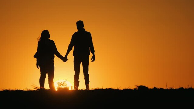 Silhouettes of a young couple holding hands and looking forward at a beautiful sunset