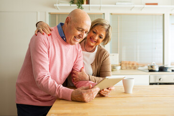 Senior couple in a kitchen, happily looking at a tablet together. The wife has her arm around her...
