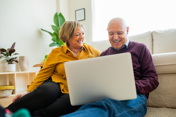 Elderly couple sitting together in a living room looking at a laptop screen. The woman stands...