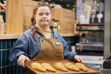 Waist up portrait of confident young woman with Down syndrome working in bakery and looking at...