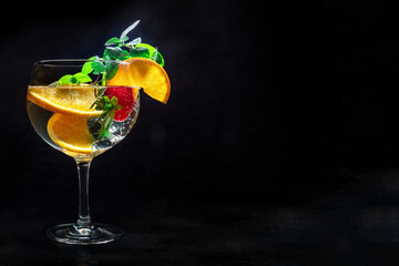 Fancy cocktail with fresh fruit. Gin and tonic drink with ice at a party, on a black background. Alcohol with orange, mint, and strawberry, with copy space