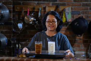 A woman was serving a glass of iced tea and a glass of hot milk; cooking equipment in the background