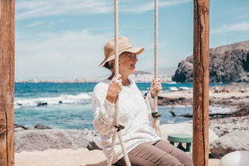 Happy senior woman sitting on swing on sea beach enjoying relaxed and carefree moments, positive retirement lifestyle for attractive senior lady wearing hat