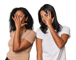 Young Latin friends in studio blink at the camera through fingers, embarrassed covering face.