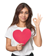 Middle Eastern woman holding paper heart cheerful and confident showing ok gesture.