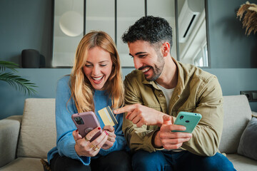 Young female holding a credit card, shopping online and her husband pointing her smart phone. Adult...