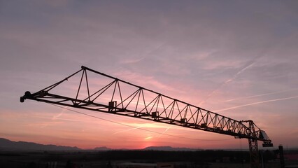 Close up during colorful sunset of Tower crane, big excavator silhouette in a construction site. Aerial drone view