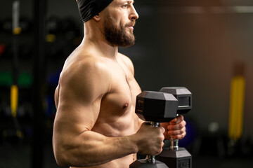 Handsome, muscular, bearded man with naked torso holding dumbbells, training in gym