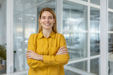 Smiling businesswoman standing with arms crossed in a modern office. She exudes confidence and...