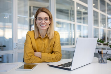 Smiling woman in a modern office setting, sitting at a desk with a laptop, wearing glasses and a yellow blouse with confidence.