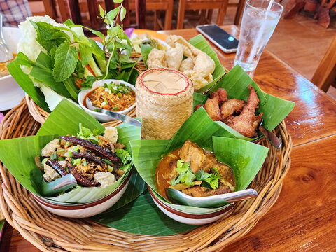 A beautifully arranged set of northern dishes served on a khantok (Lanna food container). The set meal includes various chili pastes, red curry, fresh vegetables, sticky rice, fried pork, pork caps.
