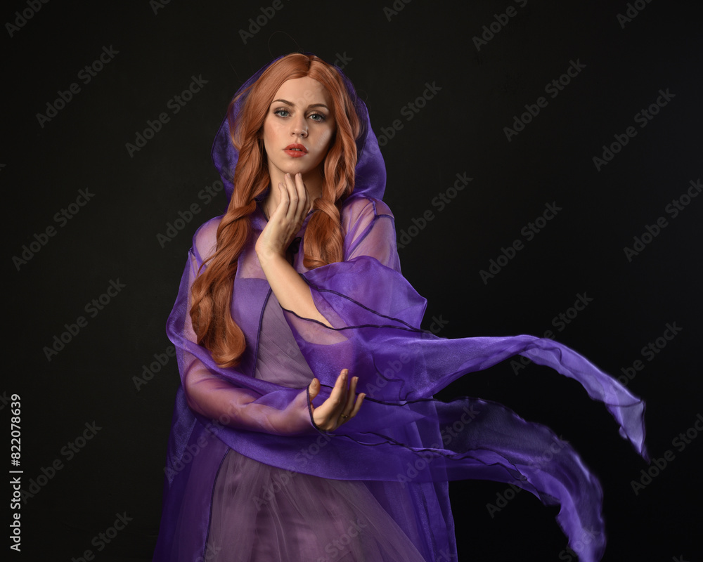 Wall mural close up portrait of beautiful female model with long red hair, wearing  purple fairy fantasy cloak gown. Elegant gestural hand poses casting magic spell. Isolated on dark black studio background