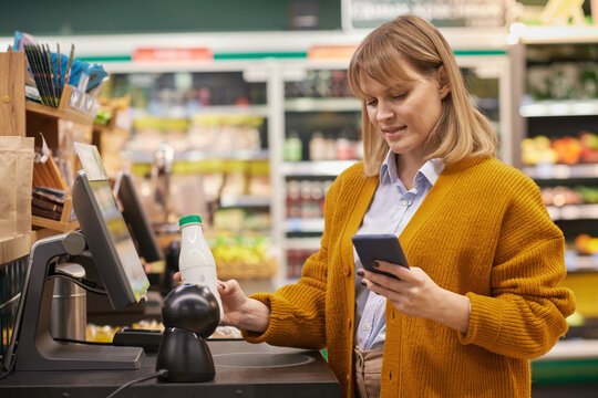 Waist up portrait of blonde adult woman scanning dairy while using self checkout service and holding phone in supermarket