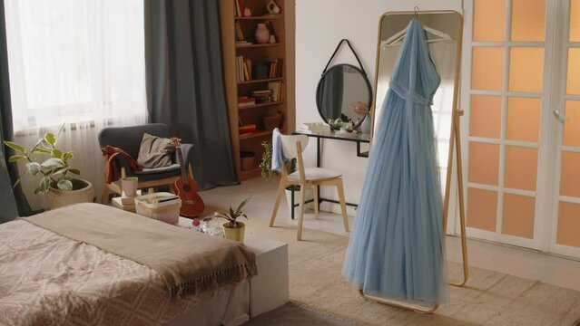 Hanger with beautiful light blue fatin evening dress hanging on large mirror standing in the center of spacious bedroom of teenage schoolgirl preparing for prom celebration