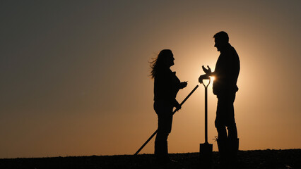 Standing in a field at sunset, a man holds a shovel while engaging in conversation with a woman,...