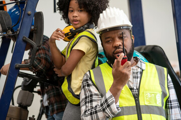 African child girl wearing a reflective vest and her father stands next to a forklift at a...