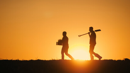 A woman farmer carries a box of seedlings, followed by a man with a shovel.