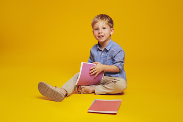 Clever little boy with blonde hair and blue shirt, sitting on studio floor and holding notebooks...