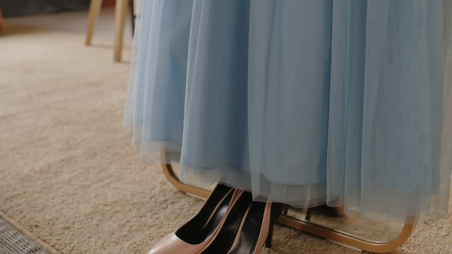 Tilt up of pair of new beige high heeled shoes standing on the floor of domestic room by mirror with long blue fatin dress prepared for prom party or some other event
