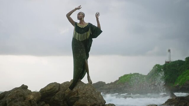 LGBTQ bipoc person in luxury dress, boutique golden jewelry poses against lighthouse on cliff, ocean, looks aside. Divine gender fluid multiethnic model in epic outfit stands on rock. Cloudy sky.