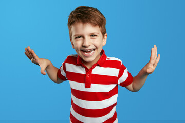 Close up portrait of excited boy in striped shirt standing with arms up on blue background while...