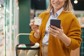 Close up of woman grocery shopping in supermarket and using smartphone in dairy aisle