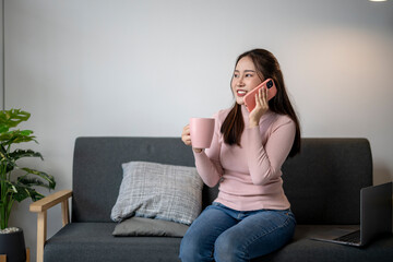 A woman is sitting on a couch and talking on her cell phone. She is holding a pink mug and a laptop