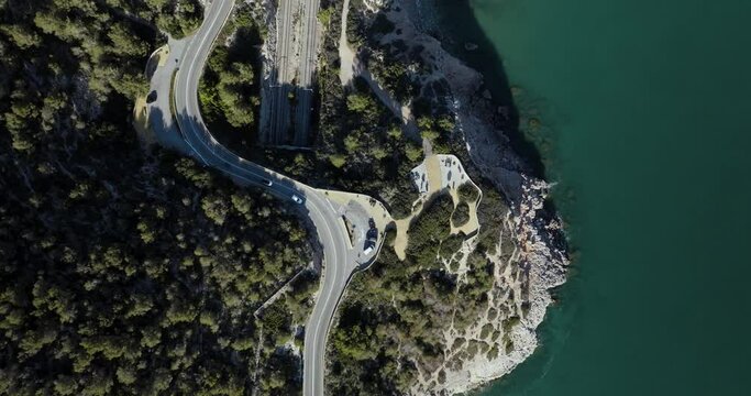 Aerial view of winding road along rocky coastline with greenery and cars, Sitges, Catalonia region, Spain.