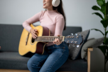 A woman is playing a guitar while sitting on a couch. She is wearing headphones and smiling. The...