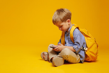 Side view of focused little boy wearing blue shirt and yellow backpack, using smartphone while...
