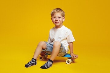 Full length of positive little boy model in stylish outfit smiling for camera and sitting on modern...
