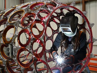 A welder wearing safety gear works meticulously on a complex, circular metal sculpture, evoking creativity, engineering, and craftsmanship