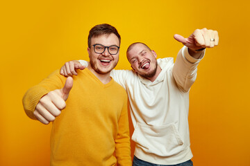 Image of two cheerful handsome guys embracing each other while showing thumbs up, wearing casual...