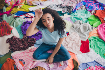 High angle view full size photo of young girl sit pile stack clutter clothes hand touch head...