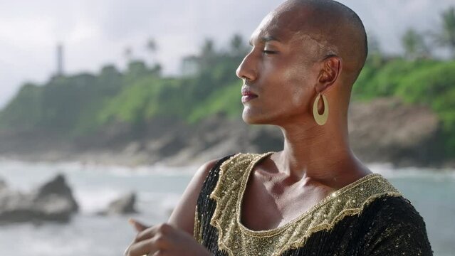 Outrageous LGBTQ bipoc person in luxury dress, boutique brass jewelry poses against lighthouse on cliff, ocean. Gender fluid multiethnic model in posh gown wears nose-ring piercing, earrings, rings.