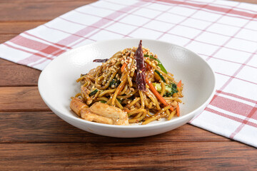 A savory plate of stir-fried noodles garnished with sesame seeds and chili peppers, served on a wooden table with a checkered napkin