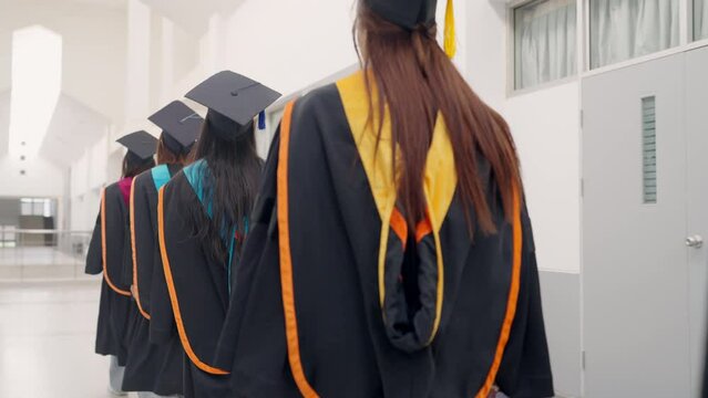 Four women in graduation gowns walk down a hallway. The gowns are black and orange, and the women are wearing caps. Scene is celebratory and happy, as the women are dressed for a special occasion