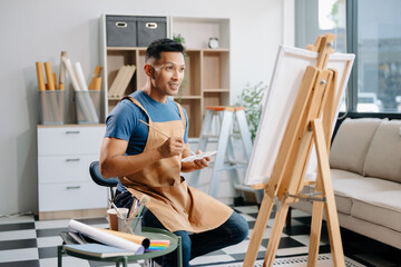 Asian male painter do artwork in art workshop, painting supplies, oil pastels, two canvas easel, creative space with paintbrush