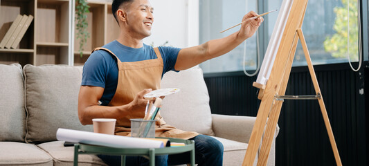 Asian male painter do artwork in art workshop, painting supplies, oil pastels, two canvas easel, creative space with paintbrush