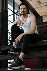 A confident man in a white tank top and black pants sits on stairs in a modern building, looking directly at the camera.