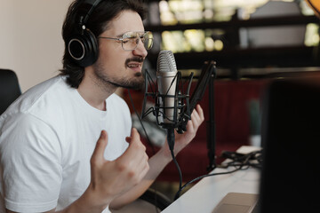 Man in a white t-shirt recording a podcast in a studio setting using a microphone and headphones.