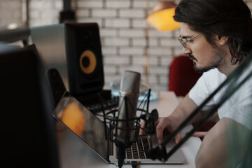 Focused man working on a laptop in a home recording studio with a microphone setup, surrounded by audio equipment.