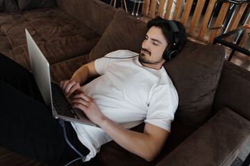 Young man lying on a sofa, using a laptop and wearing headphones, enjoying a relaxed and comfortable moment.