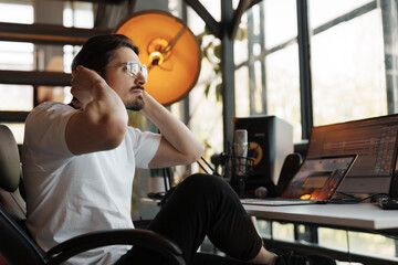 A young man taking a break and stretching while working in a modern home office with computer and equipment.