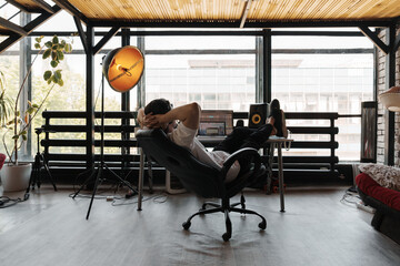 Man relaxing in a modern home office setup with large windows, natural light, and plants....