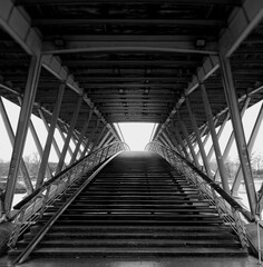Pedestrian bridge over the river. Staircase to the top. Black and white. Bridge - iron structure and wooden steps. No people. Symmetry and harmony.